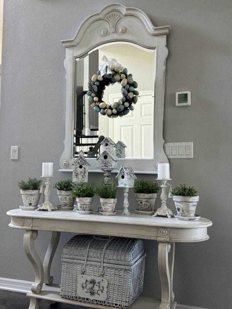 A foyer table decorated with pedestal birdhouses, plants in French flower pots and an Easter egg wreath hanging above it. 