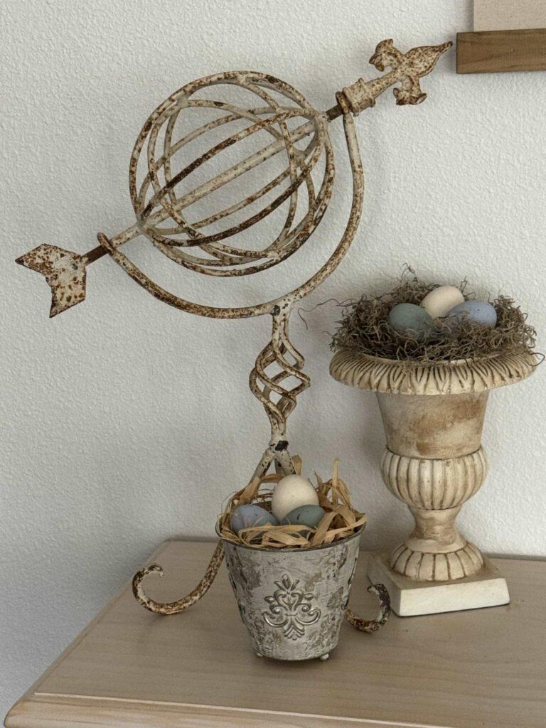 A metal garden statue, an urn filled with moss and eggs, and a metal cup filled with straw and eggs. 