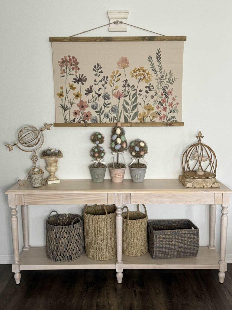 A console table with a floral canvas hanging above it. The table is decorated with three egg topiaries, a bunny under a metal cloche, a garden statue, an urn with eggs, and a metal cup with eggs. 