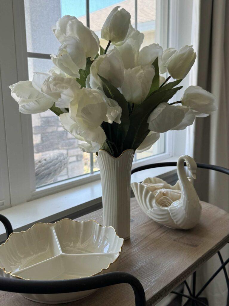 White tulips in a skinny ivory vase, next to a swan and candy dish. 