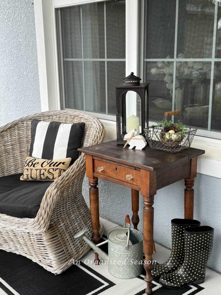 A chair, wood table, rain boots, and watering can sitting on a porch. 