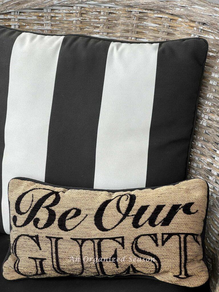 Spring porch decor idea #5 is to add throw pillows. A black and white striped one and one that says "be our guest."