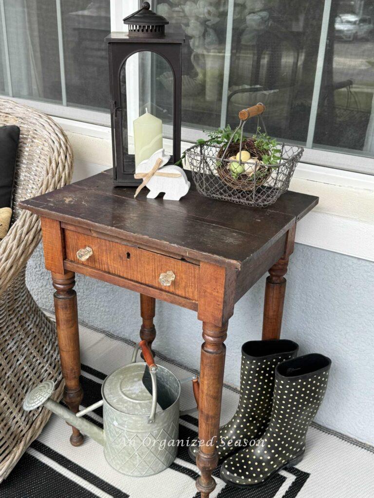 A decorated antique table with rain boots and a watering can underneath it.  