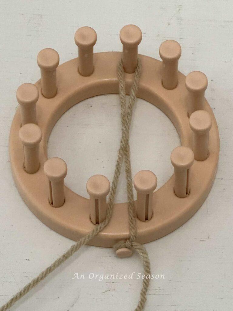 A round loom with tan yarn wrapped around one peg. 