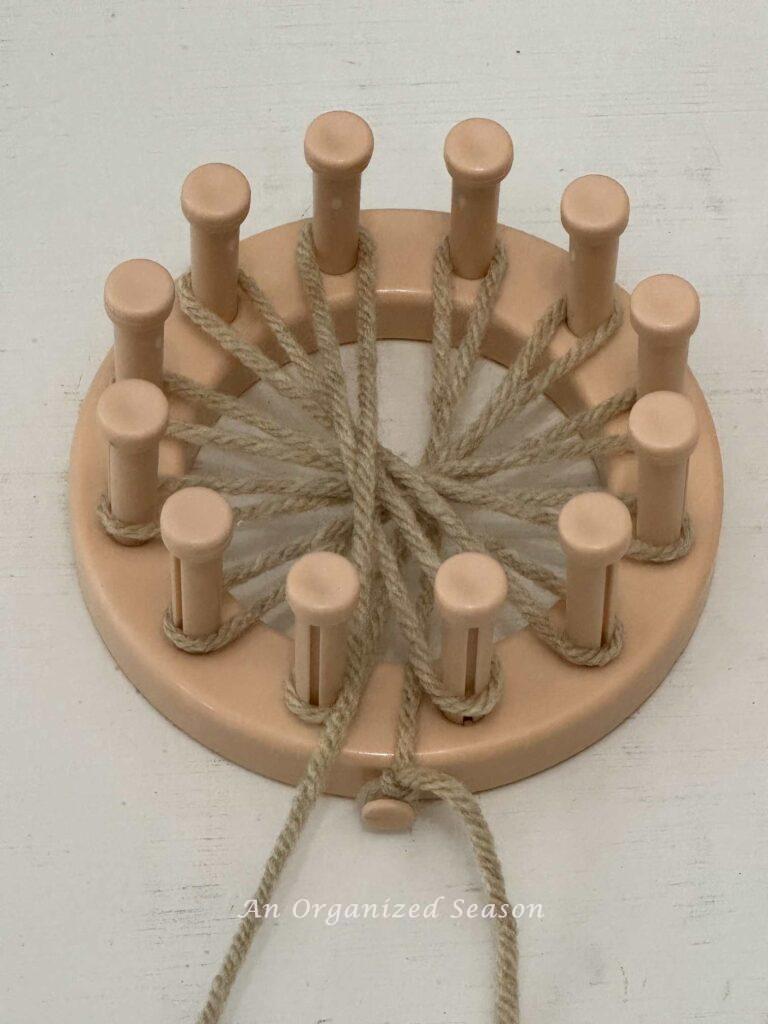 A round loom with one layer of tan yarn wrapped around all the pegs. 
