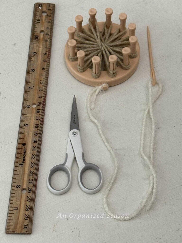 A round loom with a tan flower and white yarn attached to the anchor peg and a yarn needle. 