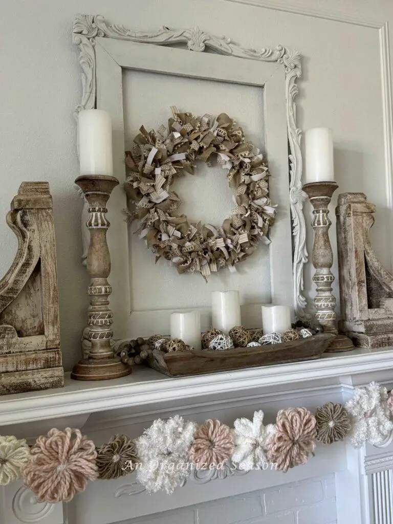 Neutral wooden decor on a fireplace mantel with ribbon wreath above and a yarn flower garland below. 