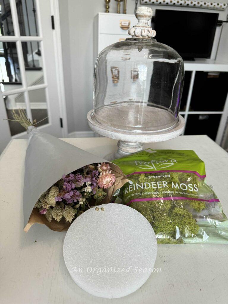 Step one to make a dried flower arrangement is to gather a cloche, dried flowers, moss, and a styrofoam disc.