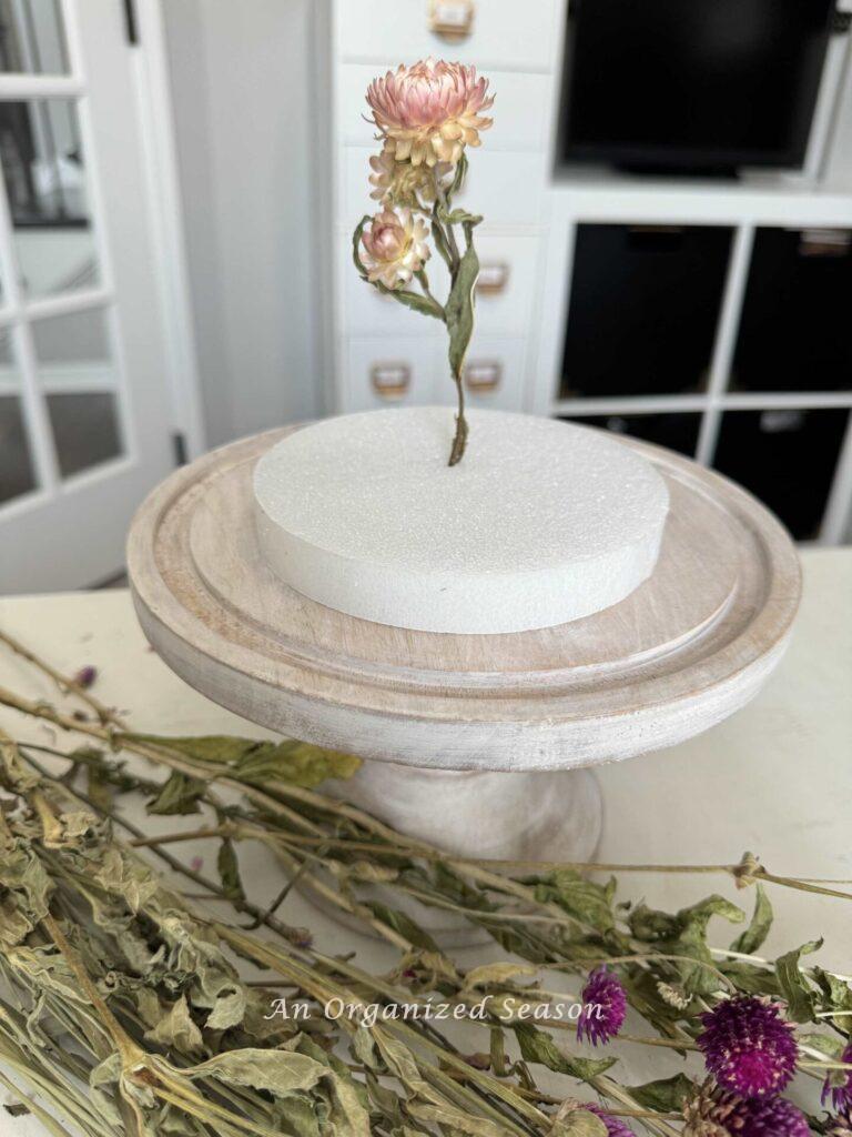 Step four to make a dried flower arrangement is to attach a flower to the center of the styrofoam base. 