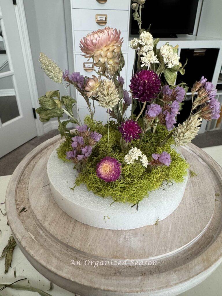 Step five to make a dried flower arrangement is to continue adding flowers and put moss around the stems. 