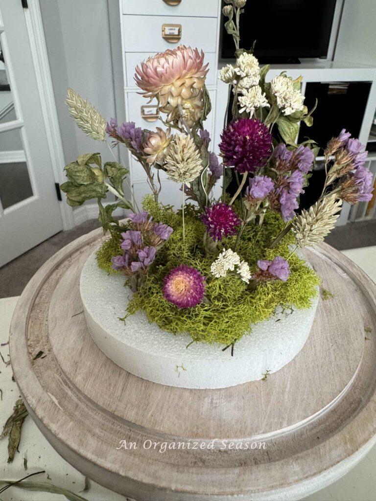 Step five to make a dried flower arrangement is to continue adding flowers and put moss around the stems.