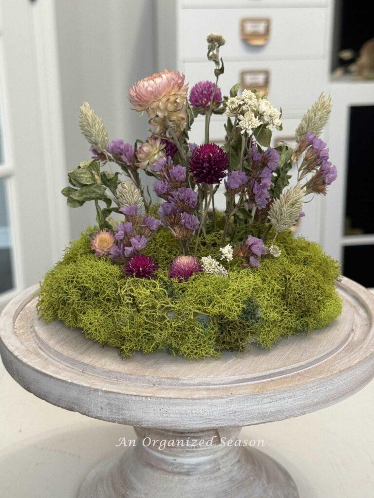 Step six to make a dried flower arrangement is to adhere moss to the sides of the base. 