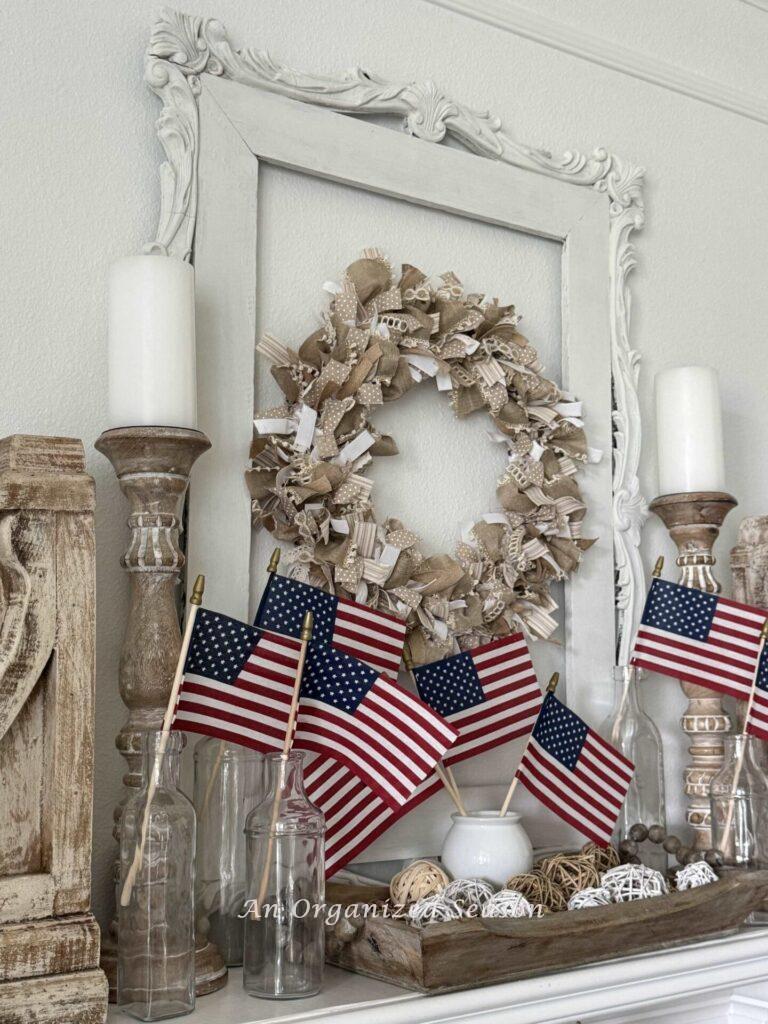 Place tall candlesticks with white pillar candles on each side of a mantel, hang a ribbon wreath, and place flags in vases across the mantel for patriotic decor.