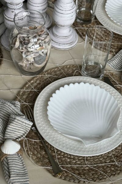 Coastal place setting with a white shell plate, napkin holder, and vase full of shells.