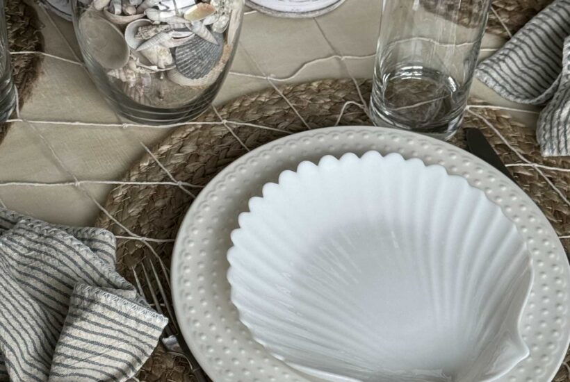 Coastal place setting with a white shell plate, napkin holder, and vase full of shells.