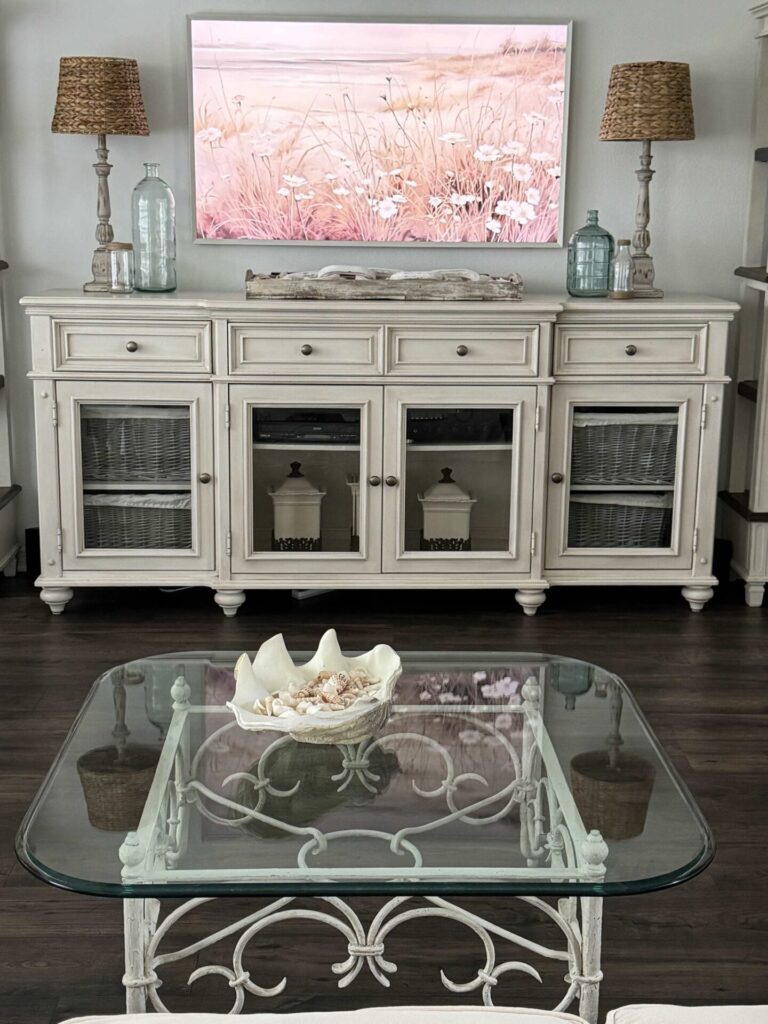 A white TV console table with two lamps, glass bottles, and a tray filled with shells. 