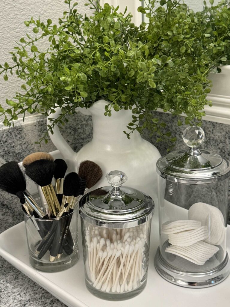 Makeup brushes in a glass next to glass canisters holding cotton swabs and pads. 