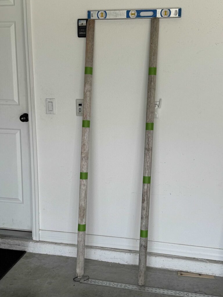 Two tree stakes propped against a garage wall with a level on top and green painters tape making where rungs should go. 