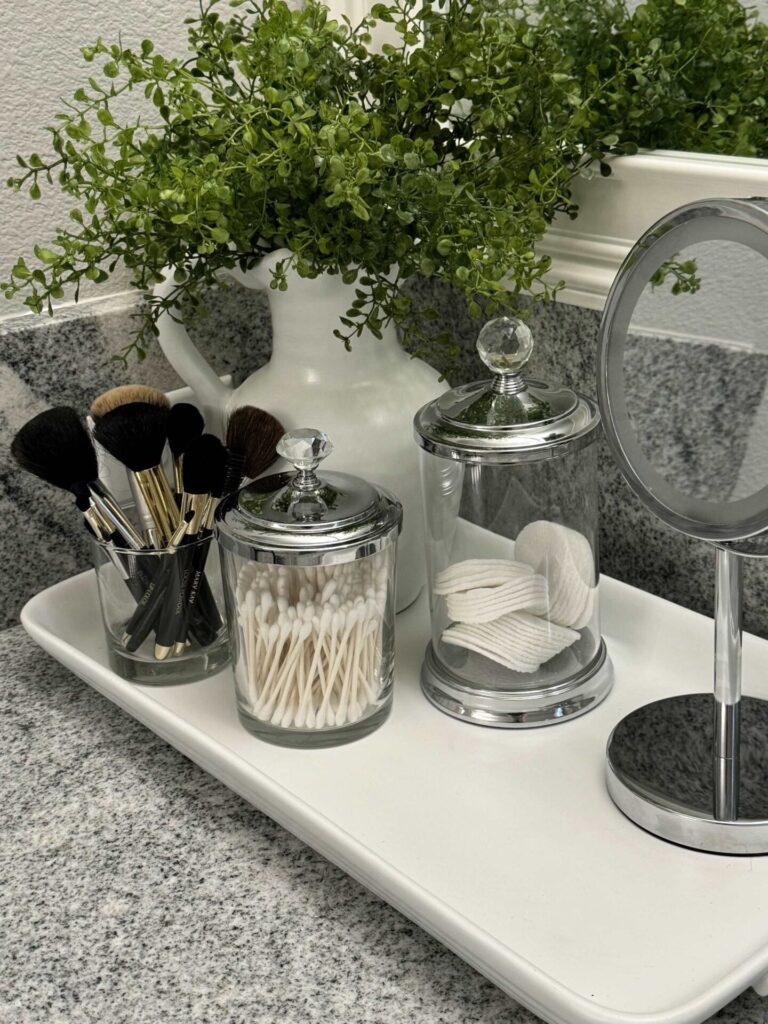 A white tray holding glass canisters, a mirror, and a plant in a white pitcher.