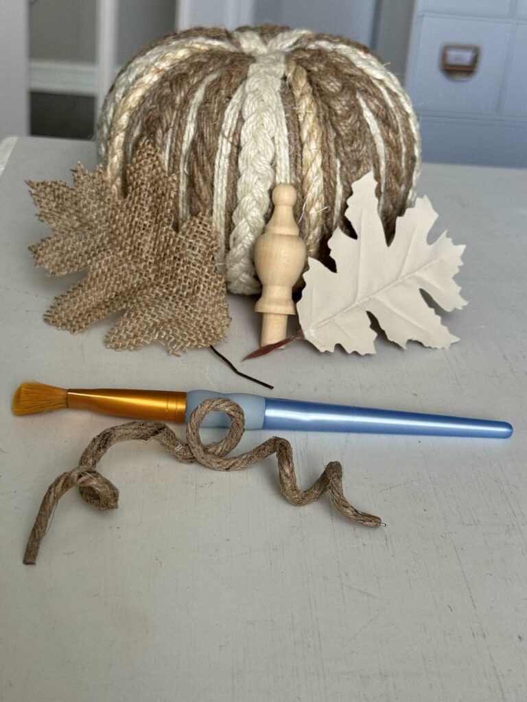 A pumpkin covered with rope and twine next to a wood finial, two faux leaves, a paintbrush, and a piece of wired jute.