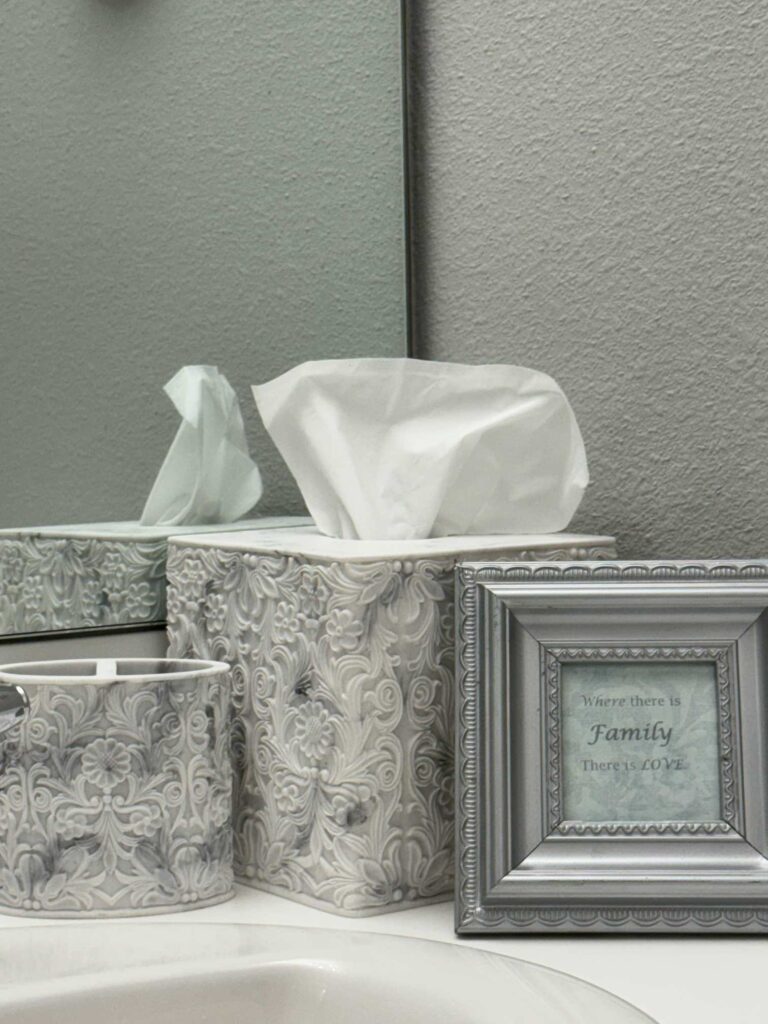 Gray and white tissue and toothbrush holder next to a small silver frame decorate a bathroom counter. 