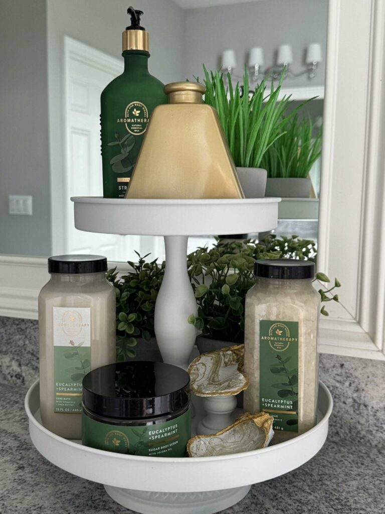 A white tiered tray holding green and gold packaged bath products. 