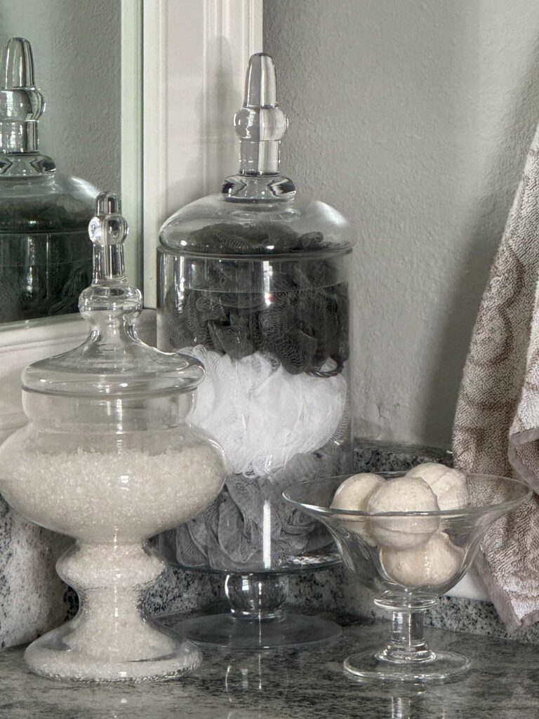 Two apothecary jars and a glass bowl  on a bathroom counter, holding bath products. 