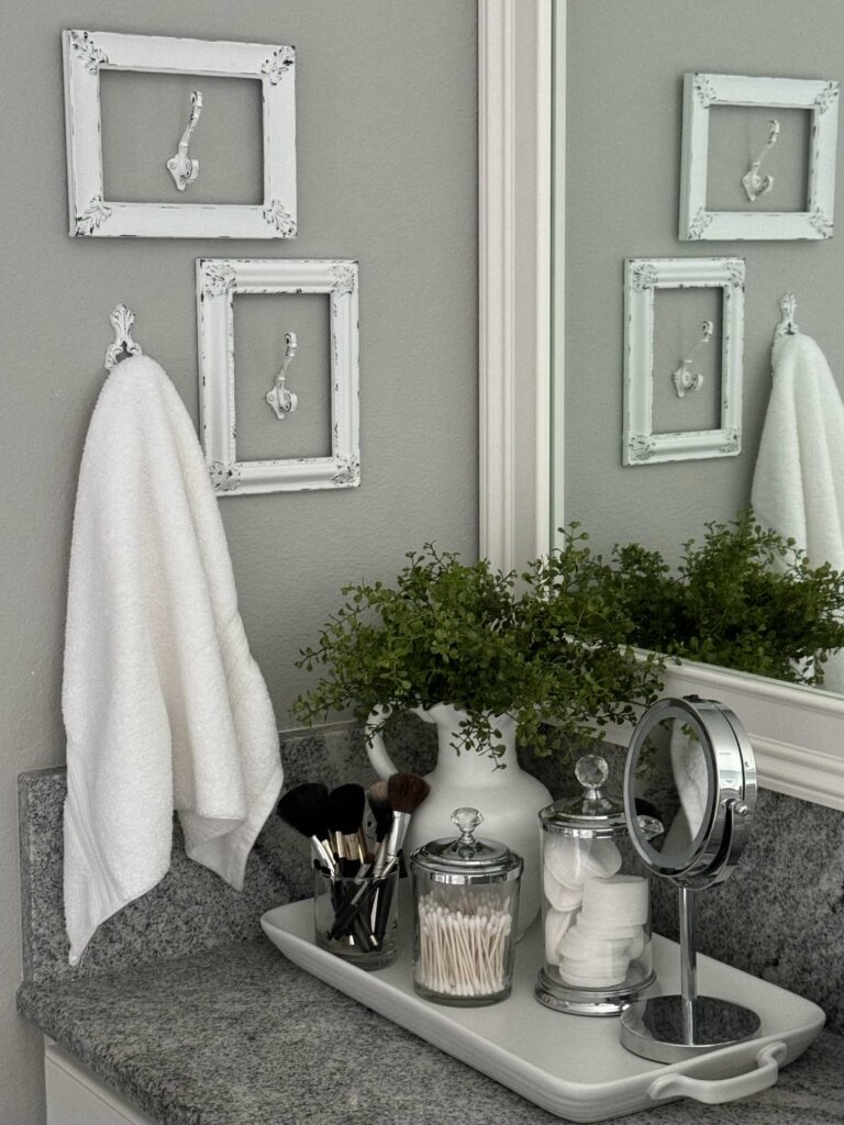 White tray holding glass canisters, a mirror, and a plant in a white pitcher, sitting on a bathroom counter. 