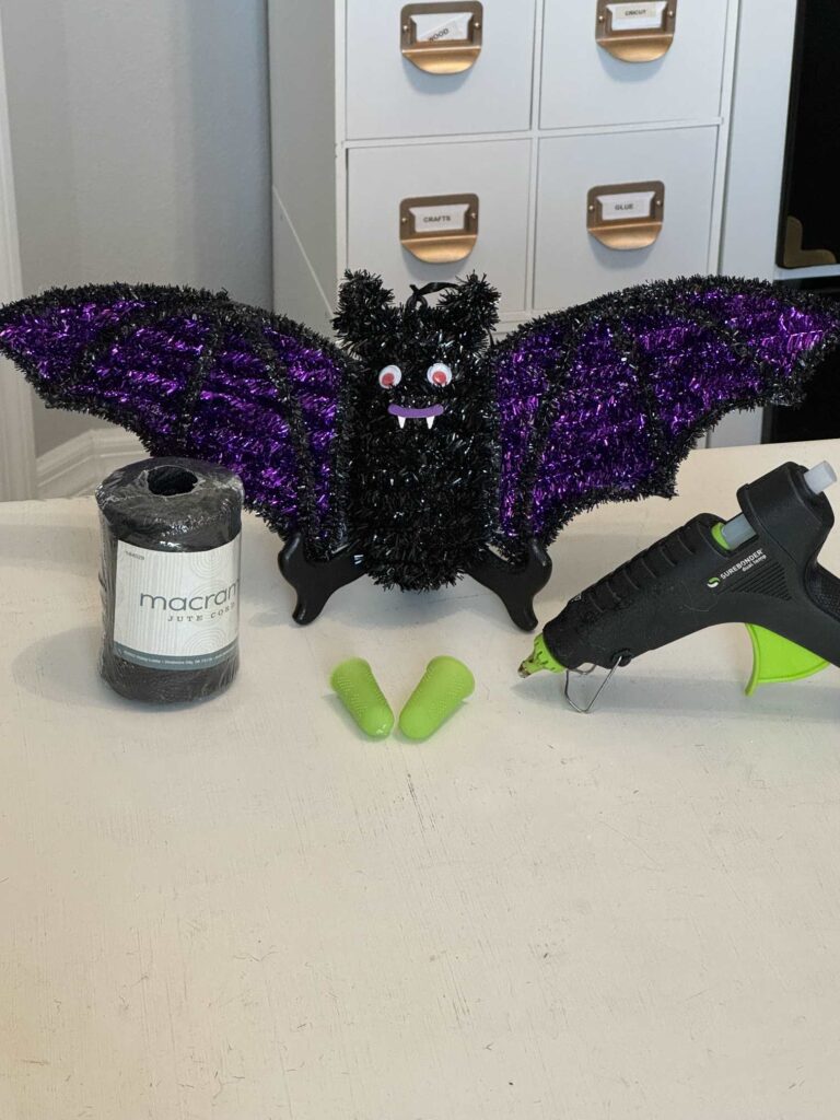 A tinsel bat, black twine, hot glue gun, and finger protectors that will be used to make a DIY Halloween bat. 