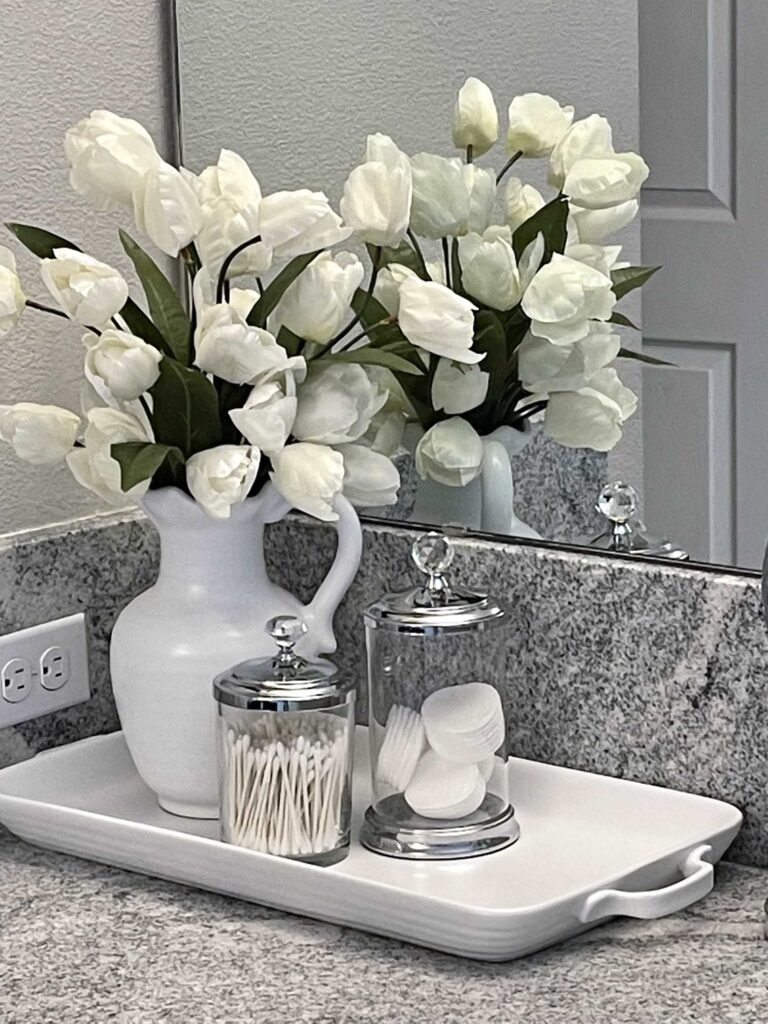 White tulips in a white pitcher, sitting on a white tray next to two glass canisters. 