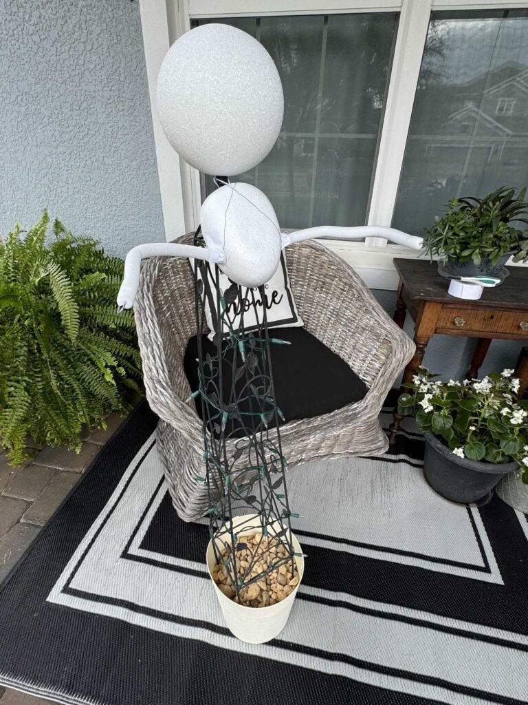 An obelisk in a planter covered with Christmas lights, with a styrofoam ball and arms attached to it. 