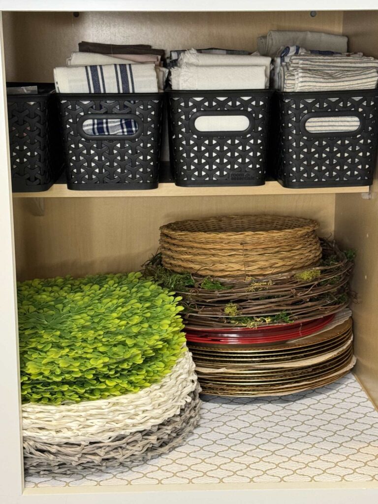 Stacked chargers and placemats and bins of napkins stored in a cabinet. 