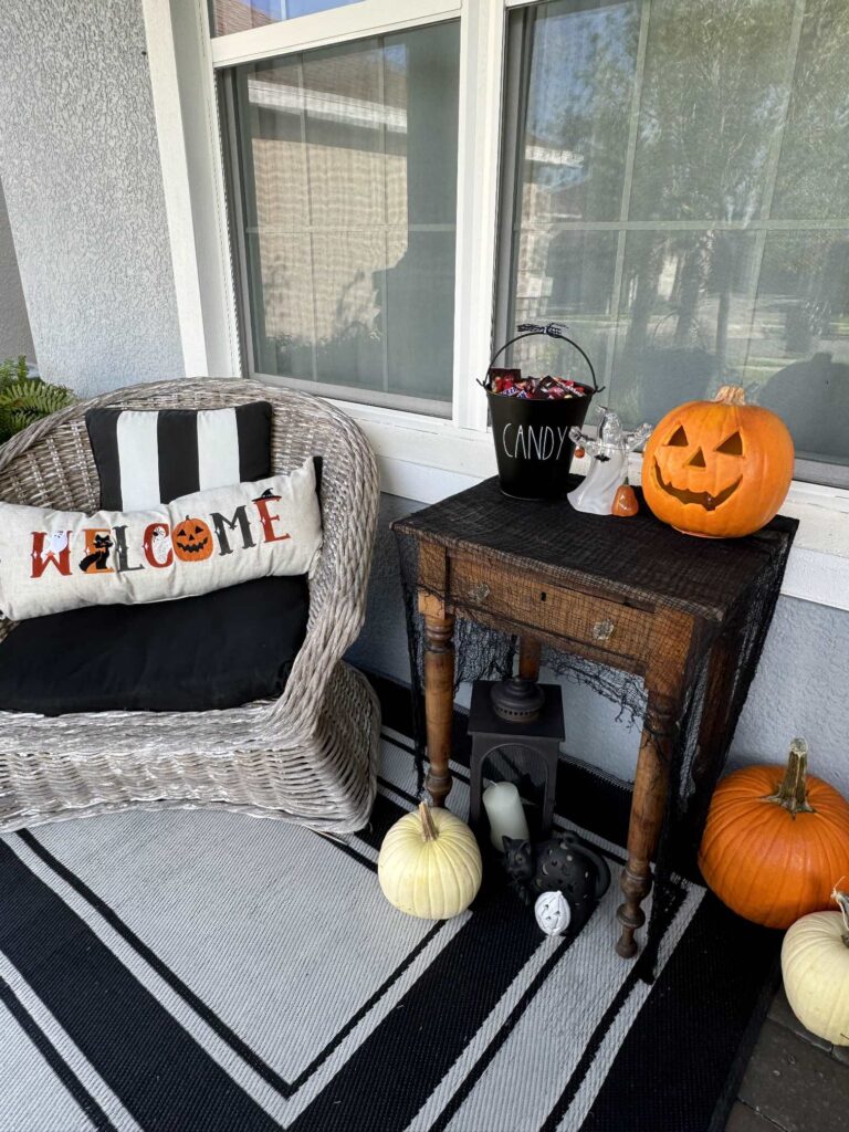 Sitting area on a porch decorated for Halloween with a jack-o-lantern, bucket of candy, and welcome pillow. 