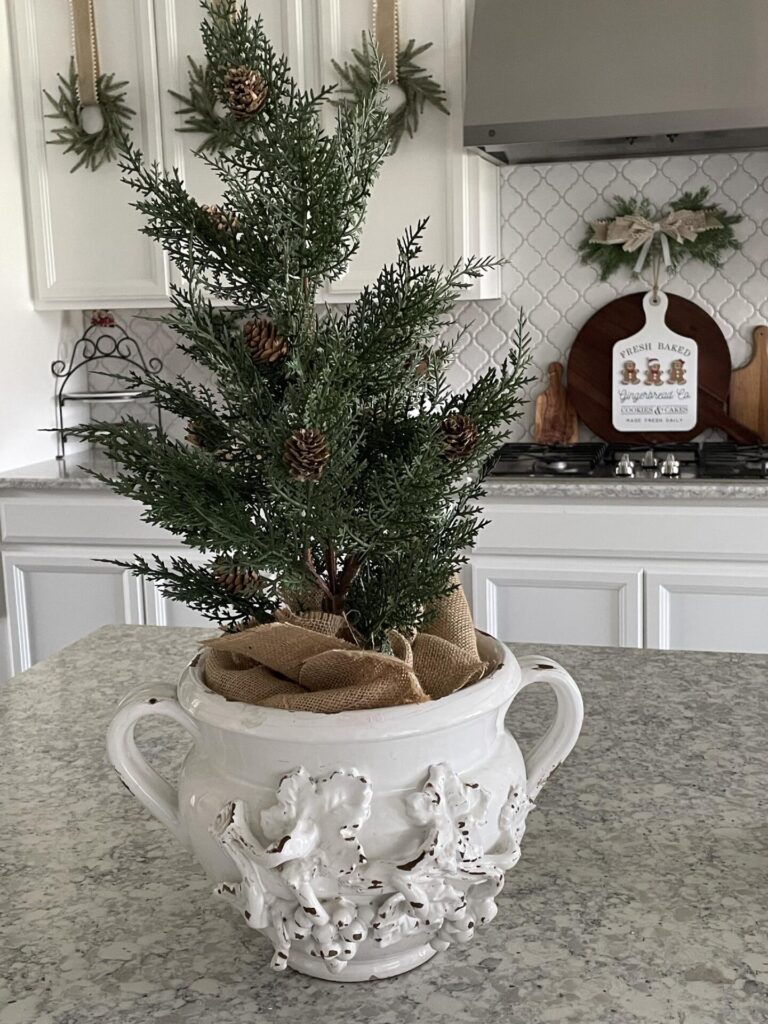 Faux evergreen tree displayed in a large white vase on a kitchen counter.