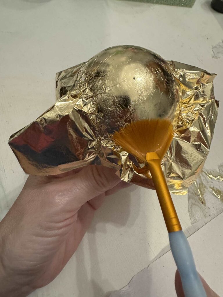 Step two for how to gold leaf is to place a sheet on the ornament and adhere it with a paintbrush. 