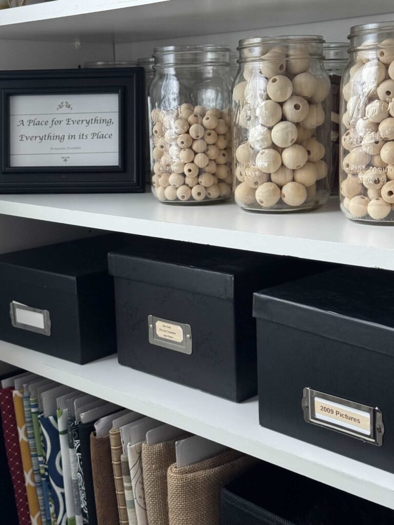 Shelves holding organized wood beads in mason jars, pictures in black photo boxes, and file-folded material pieces. 