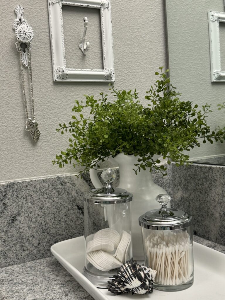 Bathroom counter tray holding white pitcher filled with greenery, two glass canisters, and a seashell.