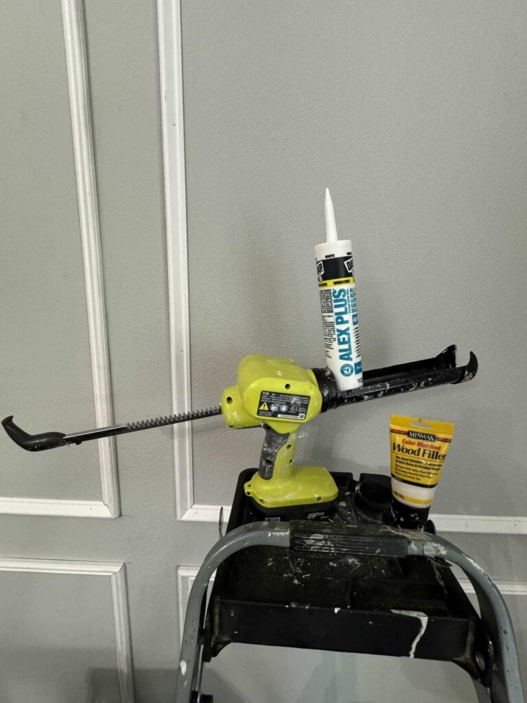 Caulk gun, caulk, and wood filler sitting on a ladder.