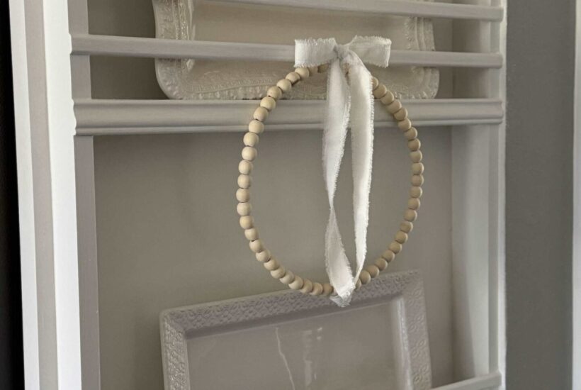 A white DIY plate rack holding white platters and a wood bead wreath with white bow.