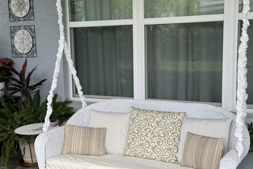 A white wicker porch swing with brown, tan. and cream throw pillows.