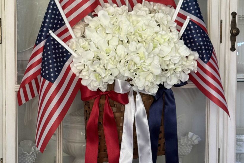 A basket wreath with flags, white hydrangeas, and red, white, and blue bows hanging on a china cabinet.