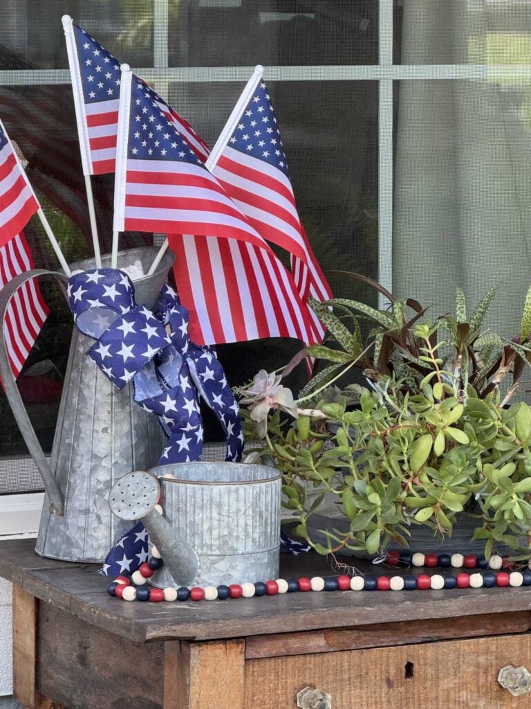 A tall metal pitcher holding American flags next to a small metal watering can, a planter of succulents, and a wood bead garland.