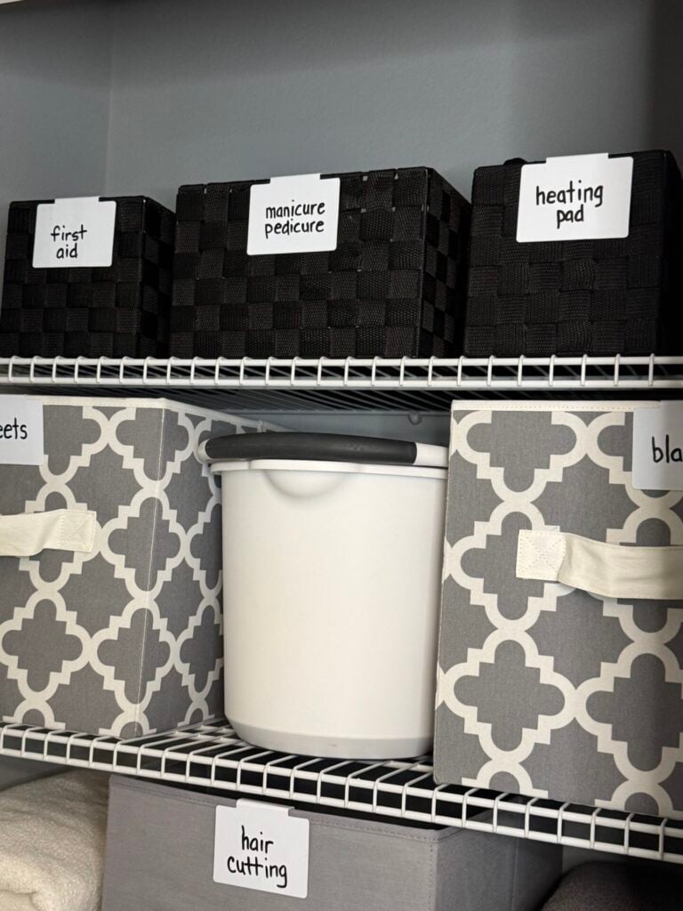 Three black woven strap baskets with white labels, a white cleaning bucket, and gray fabric bins. 