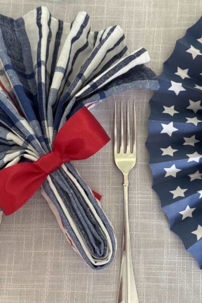A red, white, and blue napkin folded like a fan and tied with a red bow next to a blue fan with white stars.