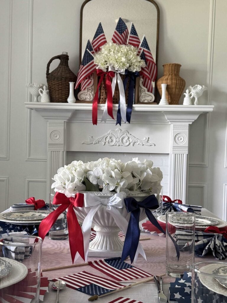 A white urn filled with white hydrangeas and red, white, and blue bows across the top sit on a table decorated for the 4th of July.