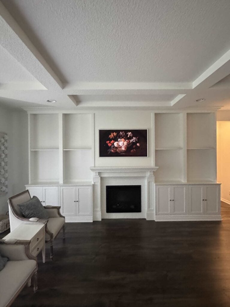 A white DIY electric fireplace and TV wall with built-in cabinets and shelves that features a frame TV with a floral arrangement and electric fireplace insert. 