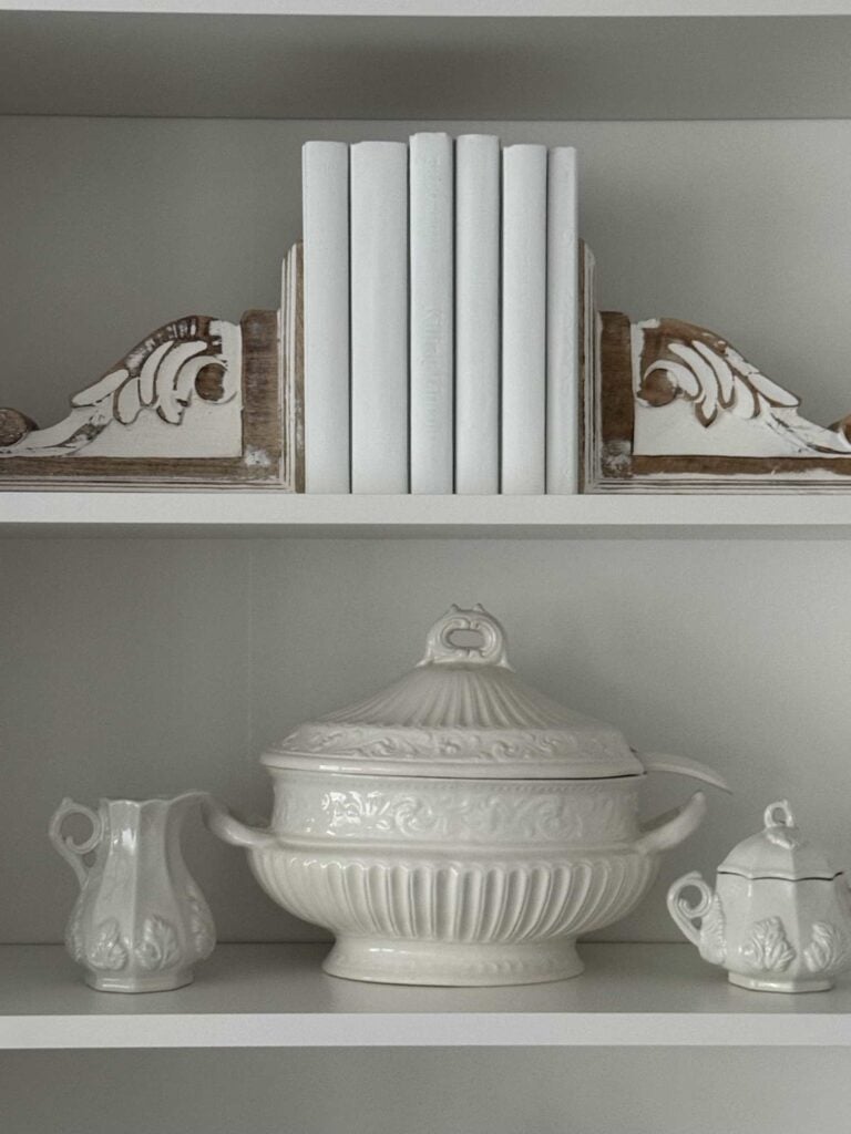 Shelves with white books held by bookends, a soup tureen, and set of ironstone cream and sugar. 