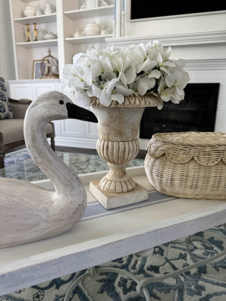 Up close pic of an urn with white hydrangeas, a wood swan, and a basket with a scalloped lid. 