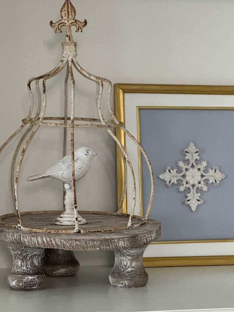 DIY artwork with gold frame, white mat, blue background, and wood embellishment, next to a metal cloche over a bird statue. 