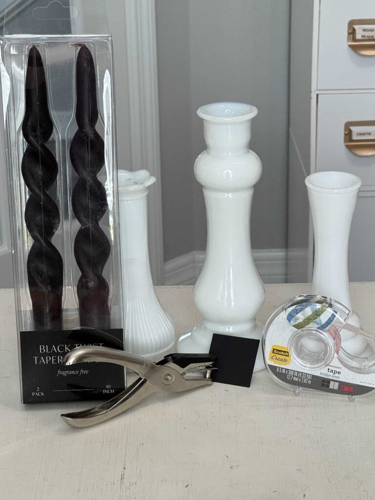 Black candles, three milk glass vases, a hole punch, black piece of cardstock, and double-sided tape. 